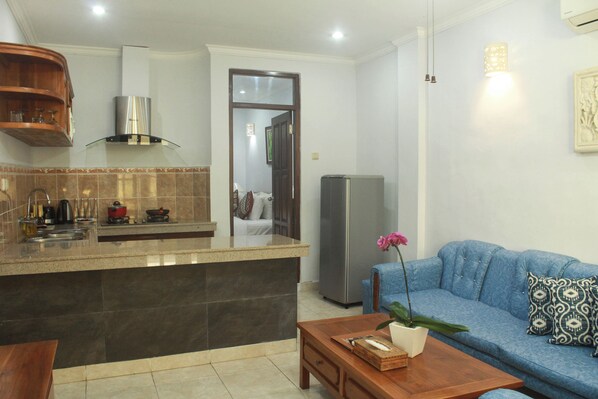 Uma sala de estar moderna com um sofá azul, uma mesa de centro de madeira e uma área de cozinha com geladeira de aço inoxidável.