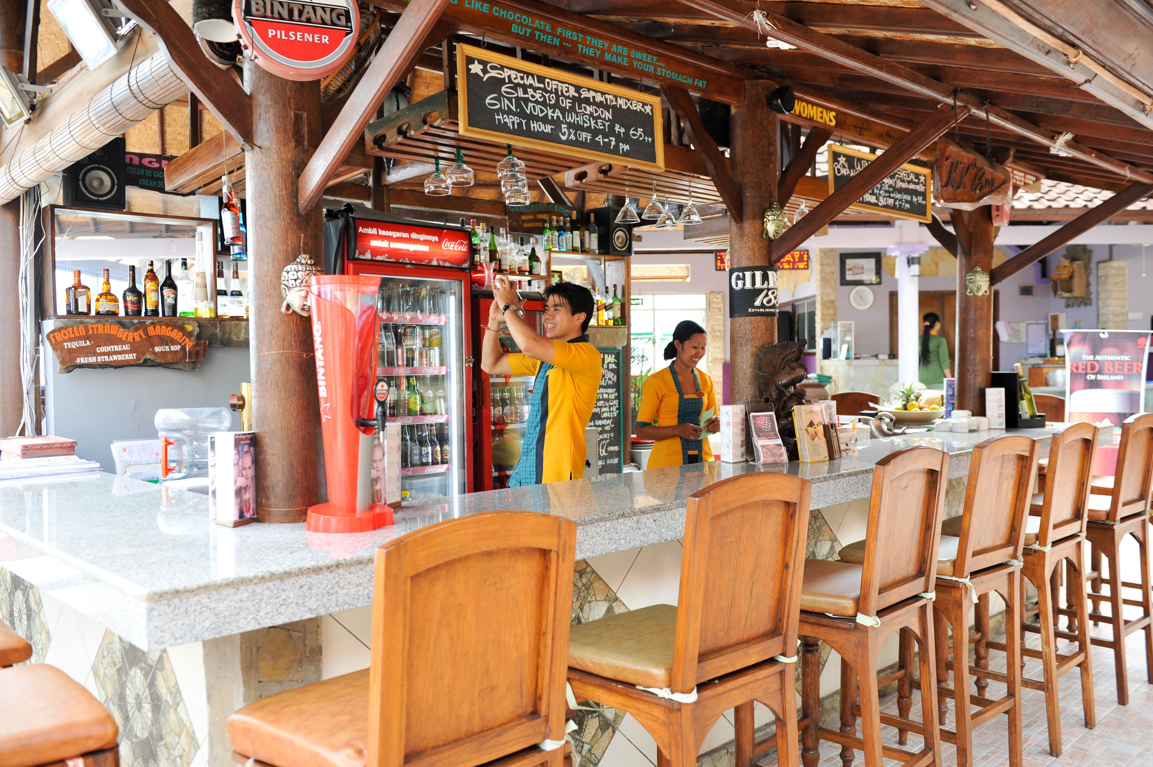 A bar area with wooden chairs and a counter. There are signs above the bar and a refrigerator with bottles.