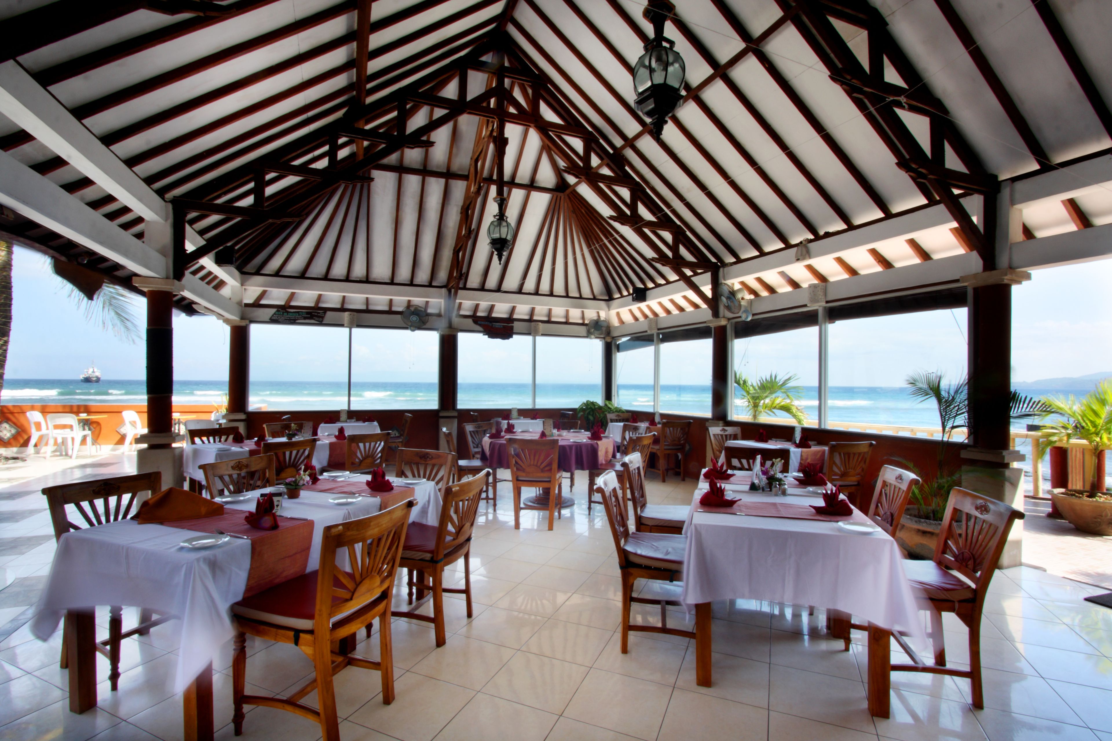 Un restaurante con mesas y sillas dispuestas para comer, amplias ventanas que ofrecen vista al océano y un techo alto con vigas de madera.