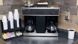 A coffee station with a Bunn VPI series espresso machine, two coffee makers, a coffee pot, and a paper cup holder with disposable cups. - The Woodridge Inn (Connersville)