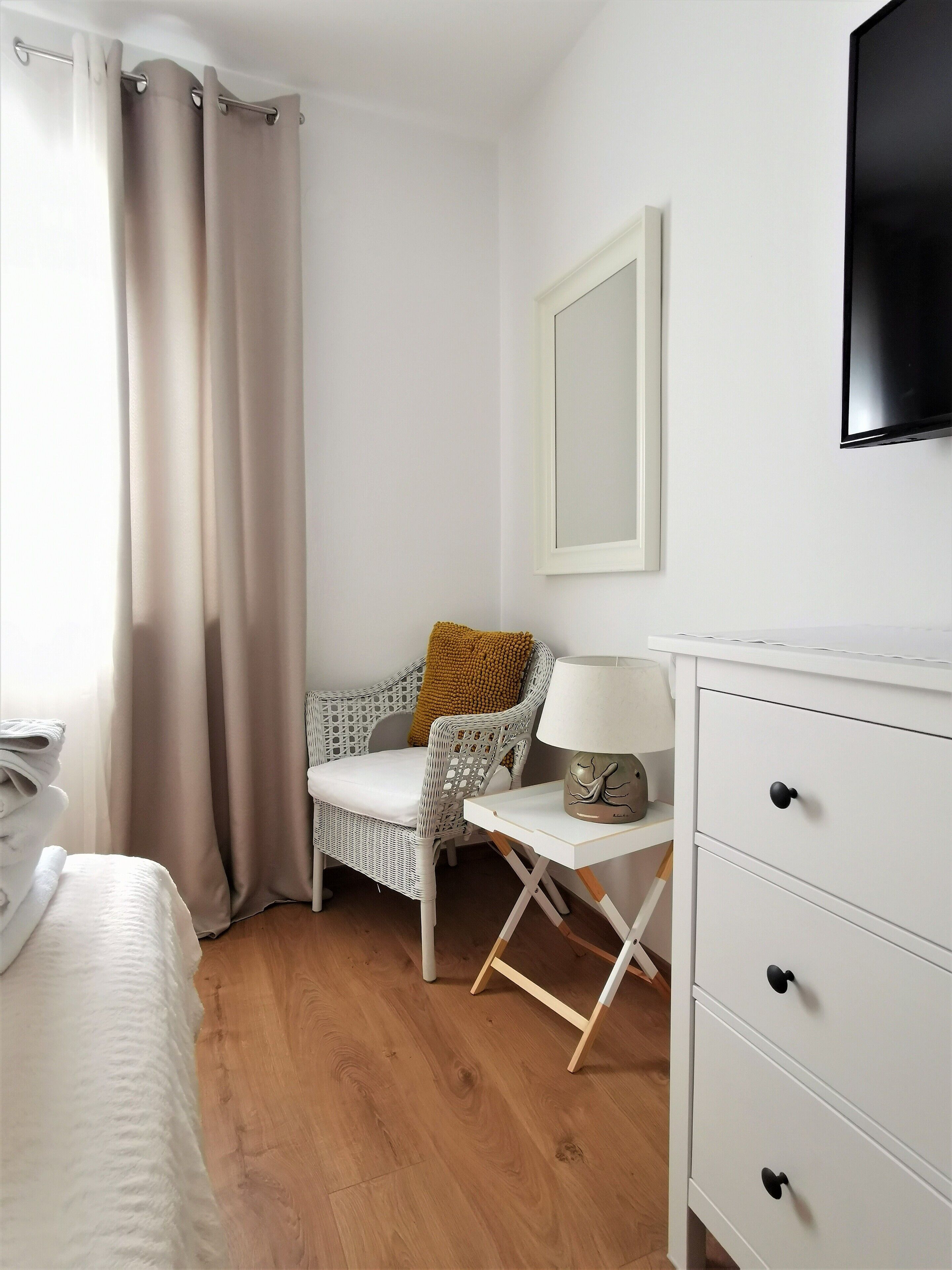 A hotel room with a bed, a chair, a table with a lamp, and a TV on the wall.