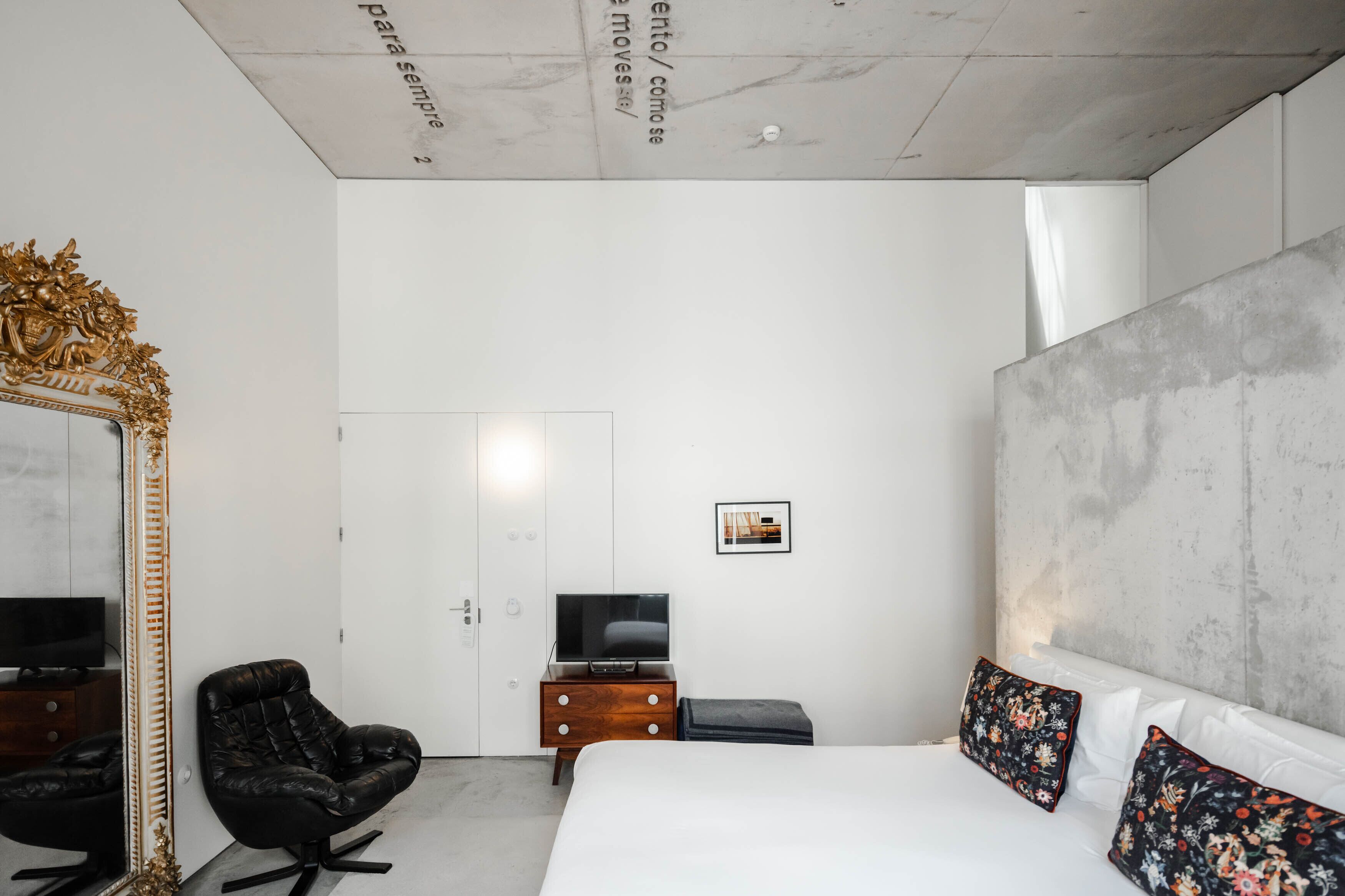 Quarto de hotel moderno com uma cama grande, uma poltrona de couro preto, uma mesa de cabeceira de madeira com uma TV, um espelho grande e uma parede de destaque em concreto.