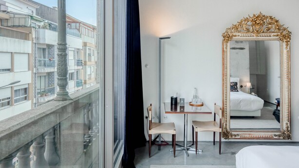 Une chambre d’hôtel avec une grande fenêtre, un lit, une table avec des chaises, un miroir et une vue sur les bâtiments environnants.