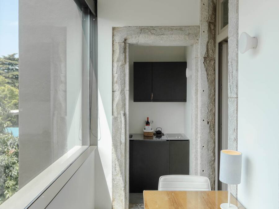 Ein Balkon mit Holztisch und weißem Stuhl, eine Küchenzeile mit Schränken und Blick ins Grüne.