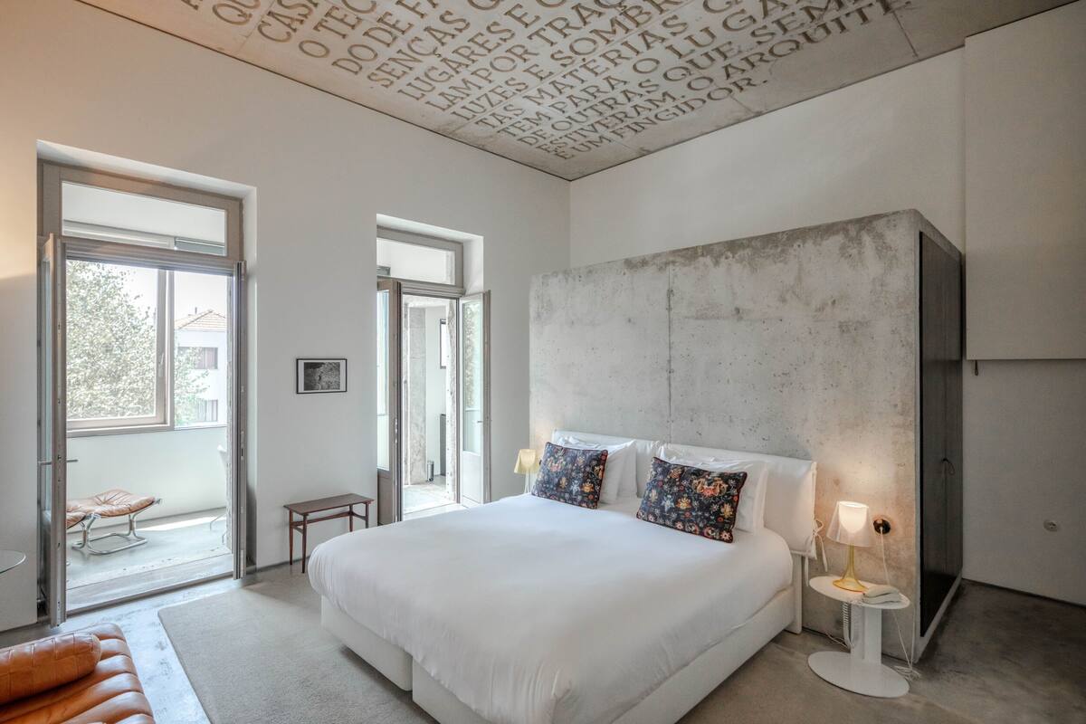 A modern bedroom with a large bed, a small bedside table, a nightstand, and a window with a view of the outside.
