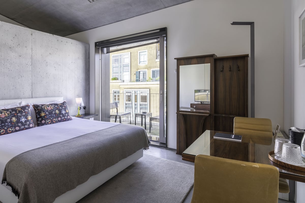 A modern hotel room with a large bed, a desk, and a view of the city through a large window.
