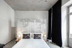 Una habitación de hotel moderna con una cama grande, dos almohadones decorativos, una mesita de noche con lámpara y vistas al exterior a través de una ventana con cortinas negras.
