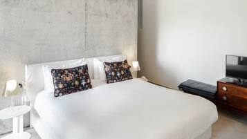 Un dormitorio moderno con una cama grande, dos almohadas, una mesita de noche con lámpara y un televisor sobre un aparador de madera.