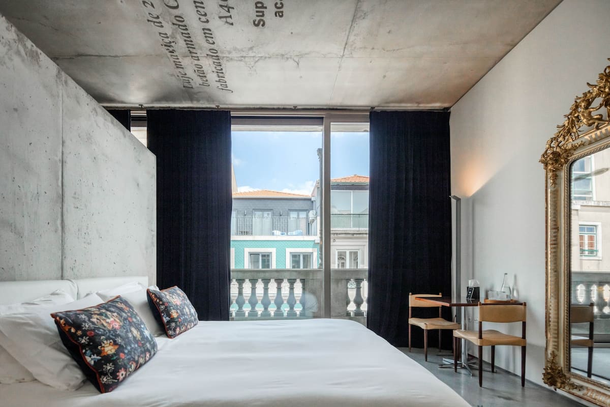 A bedroom with a large bed, two decorative pillows, a view of buildings through a balcony, and a large mirror.