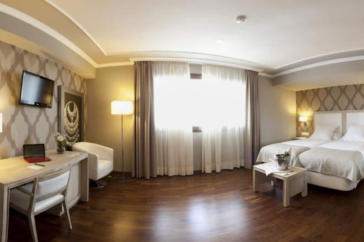 Habitación de hotel con escritorio, dos camas, televisor y un amplio ventanal con cortinas.