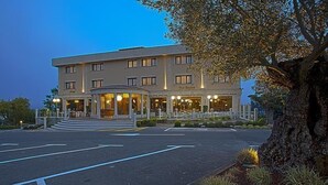 Un hotel de varios pisos con una entrada central, un área ajardinada con un árbol grande y un estacionamiento.