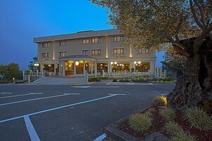 Un hotel de varios pisos con una entrada central, un área ajardinada con un árbol grande y un estacionamiento.