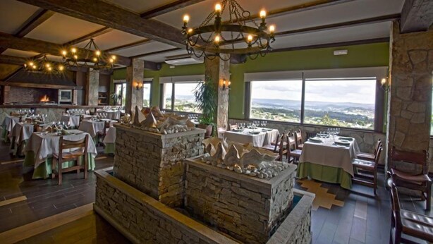 Un restaurante con chimeneas de piedra, vigas de madera y ventanas amplias que ofrecen una vista. Mesas listas para comer.
