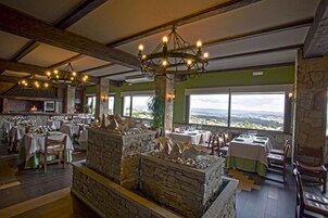 Un restaurante con chimeneas de piedra, vigas de madera y ventanas amplias que ofrecen una vista. Mesas listas para comer.