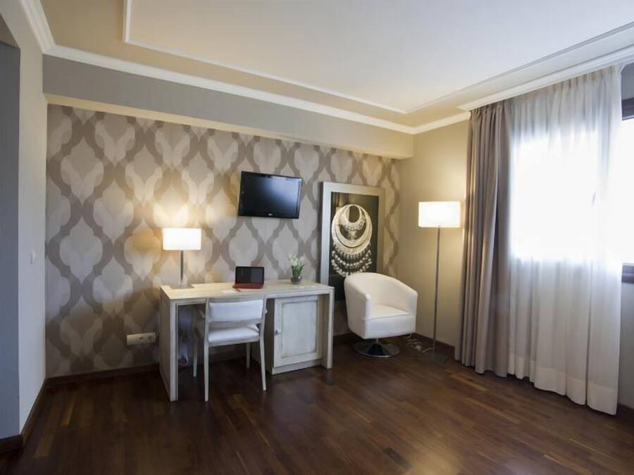 A hotel room with a wooden floor, a desk with a chair, a television mounted on the wall, and a large window with curtains.