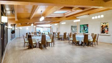 A spacious dining area with high wooden ceilings and natural light from skylights. Tables are set with chairs, and artwork is displayed on the walls.