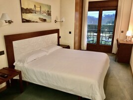 Una habitación de hotel con una cama grande, dos mesitas de noche, un cuadro de un puente, una ventana con vistas a un balcón y una puerta.