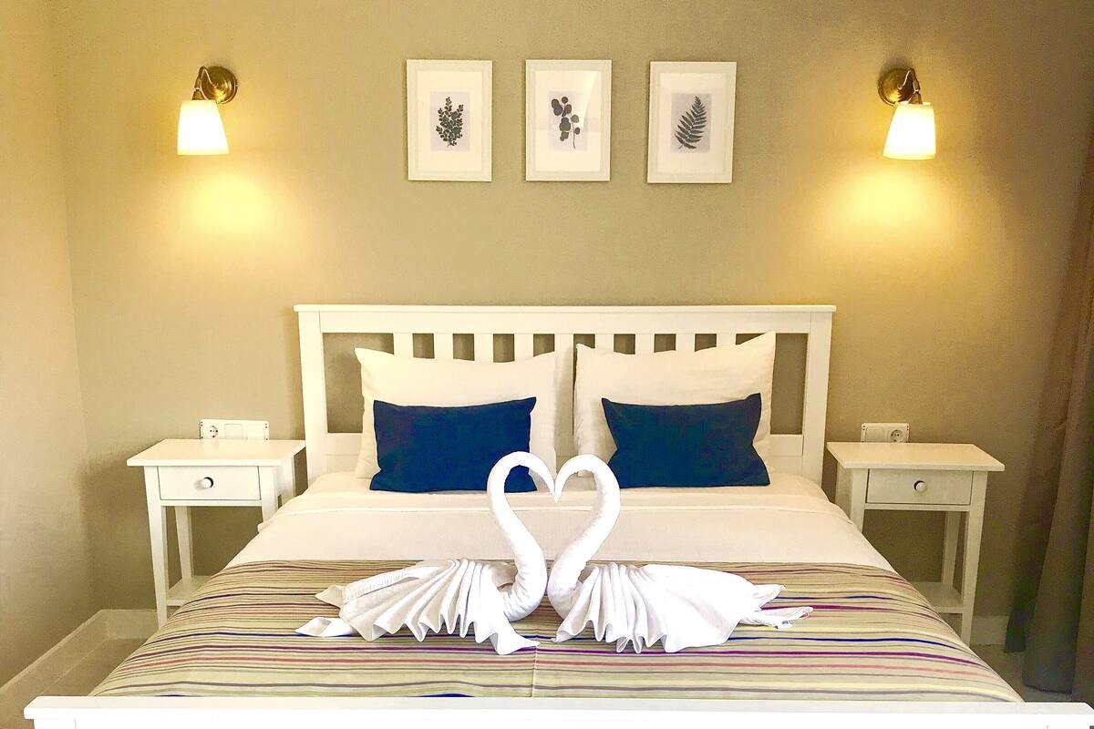 A neatly made bed with a heart-shaped towel decoration, two bedside tables, and wall-mounted light fixtures.