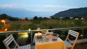 Uma mesa posta para refeição, com vista para o mar e colinas ao fundo.