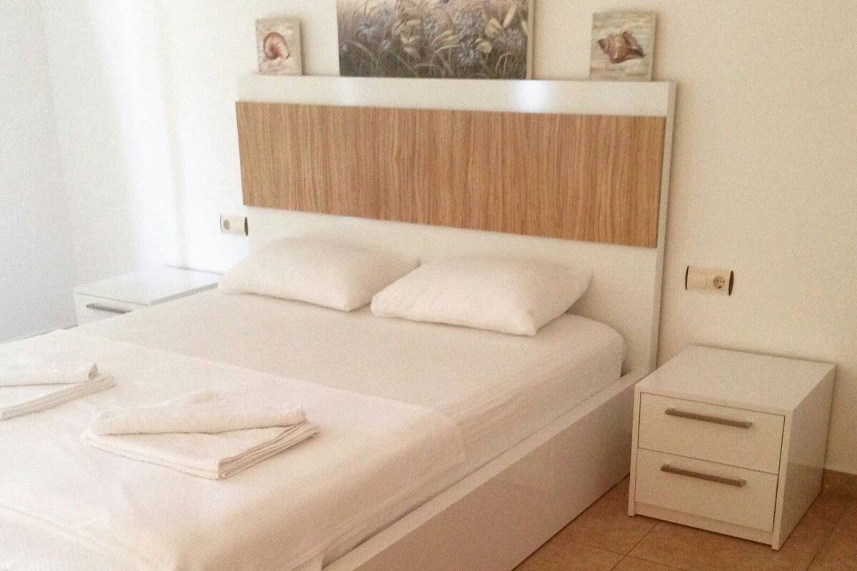 A neatly made bed with pillows and a folded towel, a bedside table with drawers, and a nightstand with a lamp.