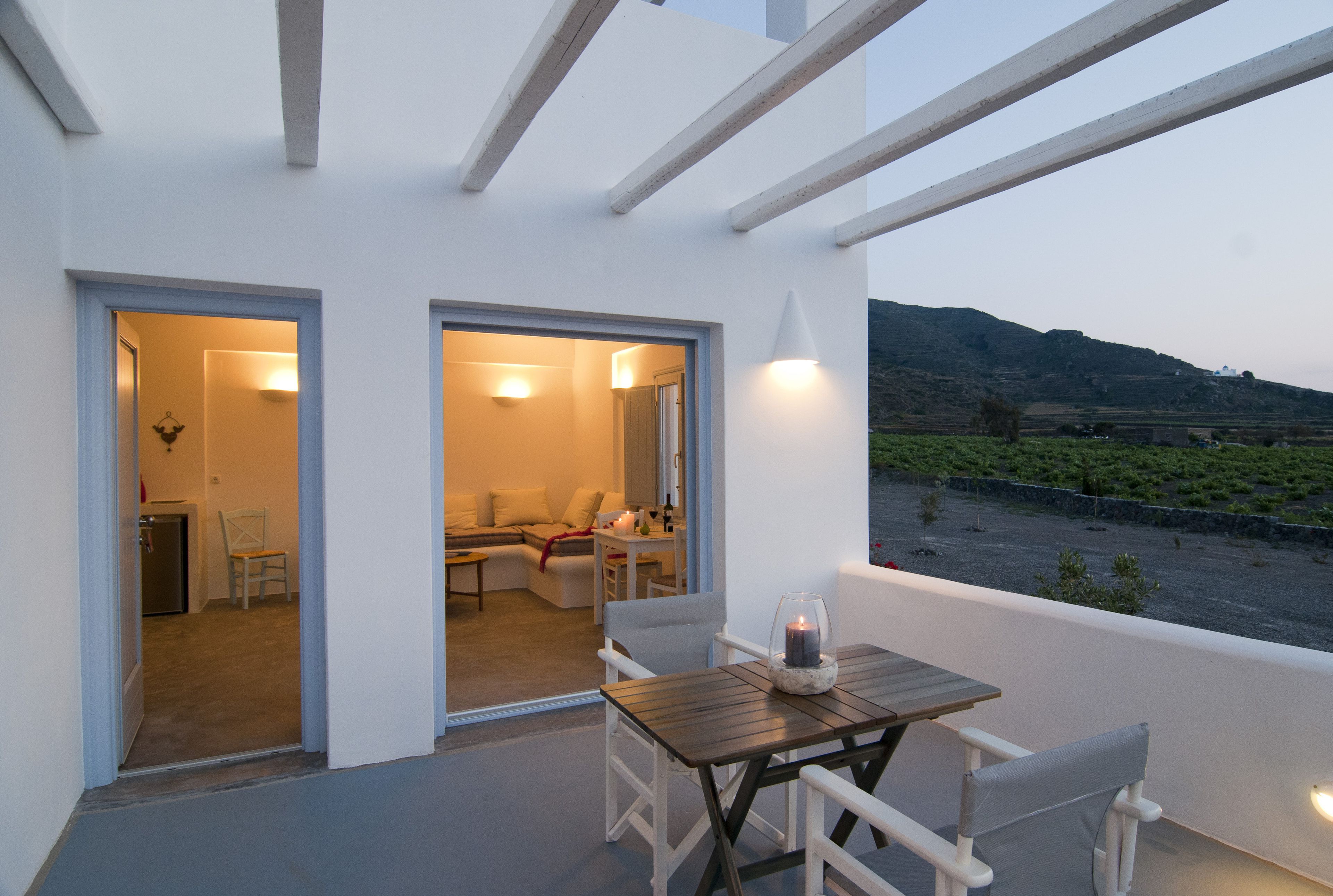 A balcony with a table and chairs, a view of a vineyard, and a building with an open door leading to a living area.
