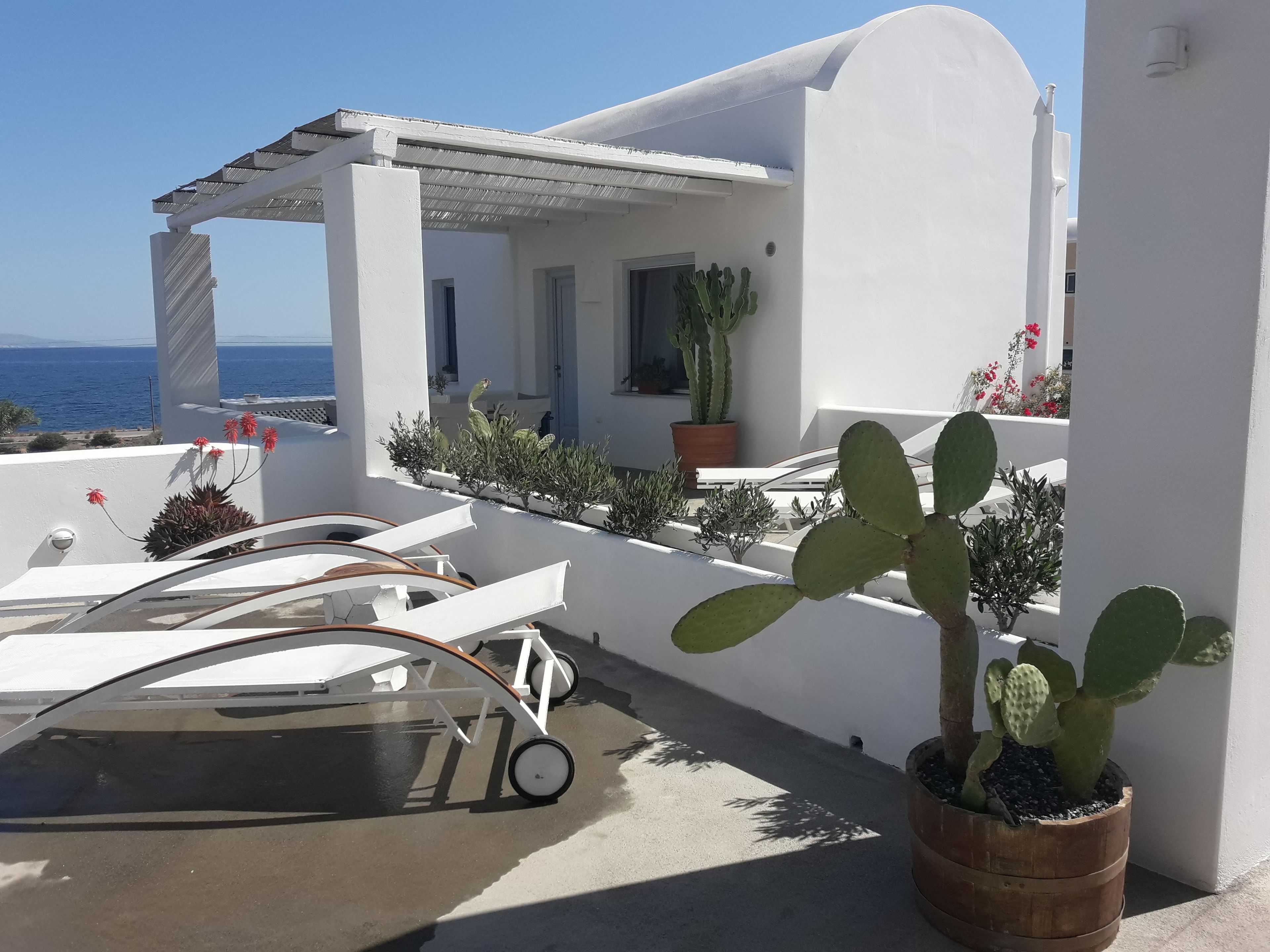 A white building with a balcony overlooking the sea. There are potted plants and a small table with chairs on the balcony.