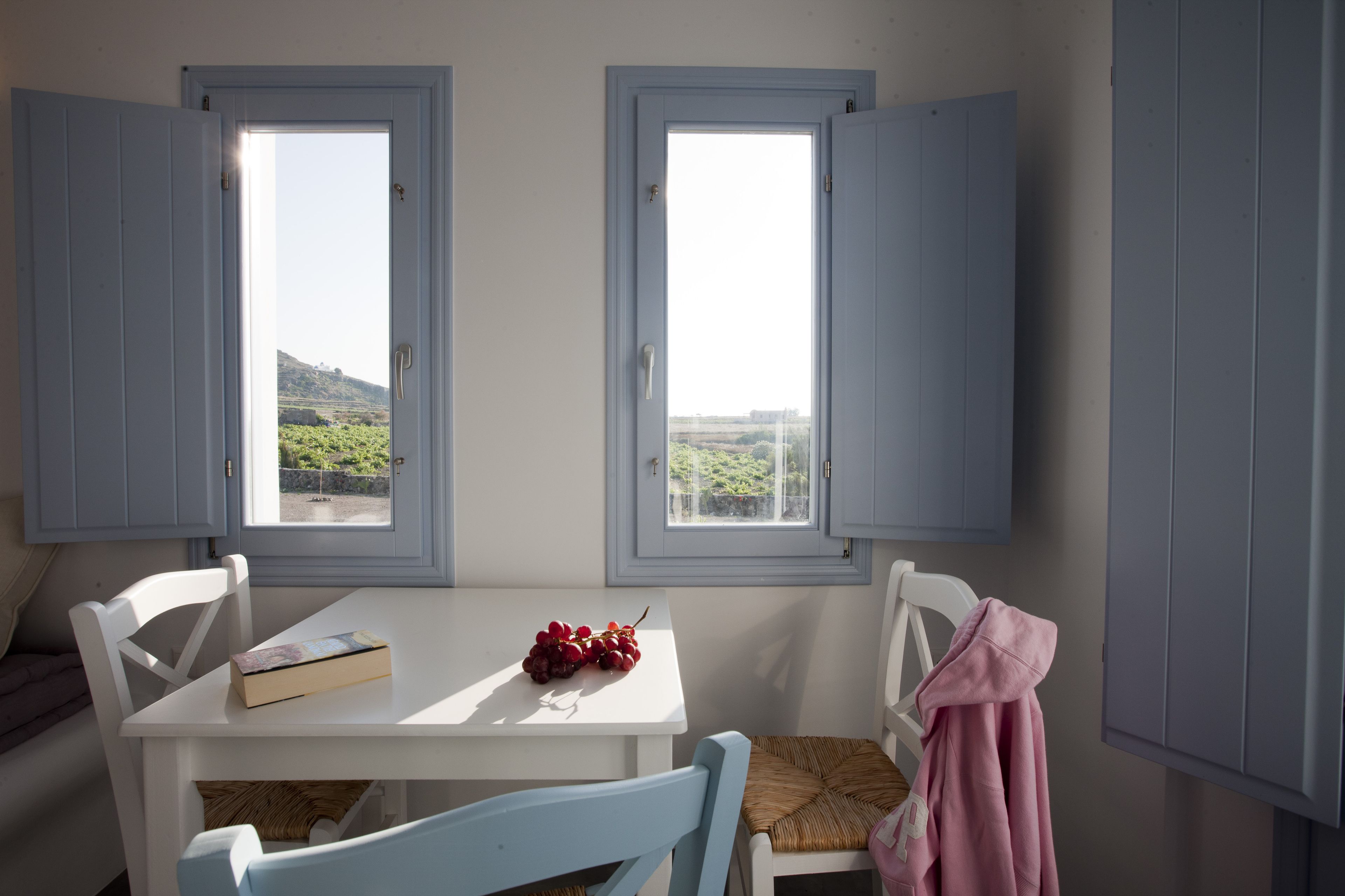 Une pièce avec deux fenêtres, une table sur laquelle se trouvent un livre et des raisins, et une chaise sur laquelle est posé un objet rose.