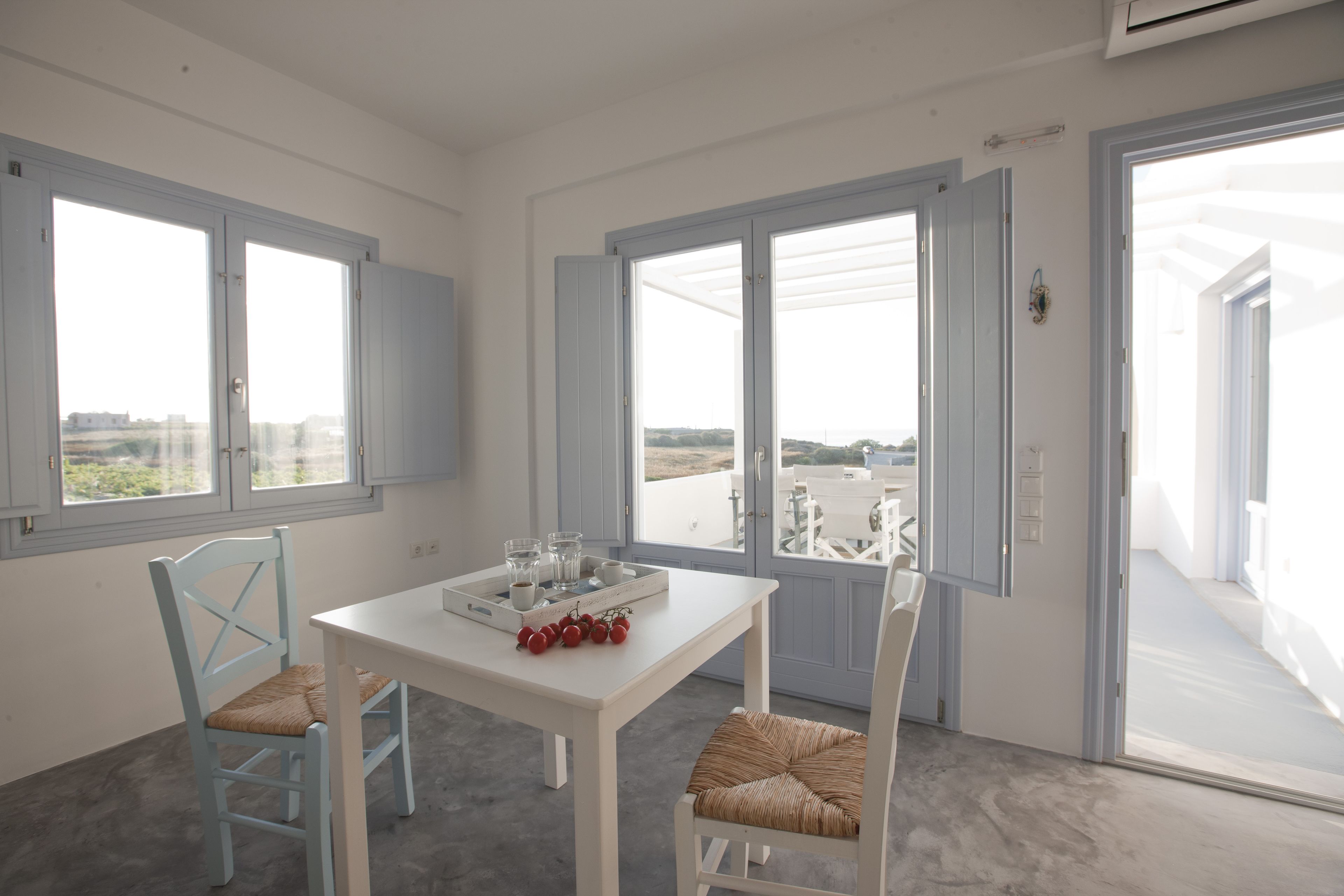Un espace repas moderne avec une table et des chaises blanches, un plateau contenant des fruits et des verres, et de grandes fenêtres offrant une vue sur la ville.