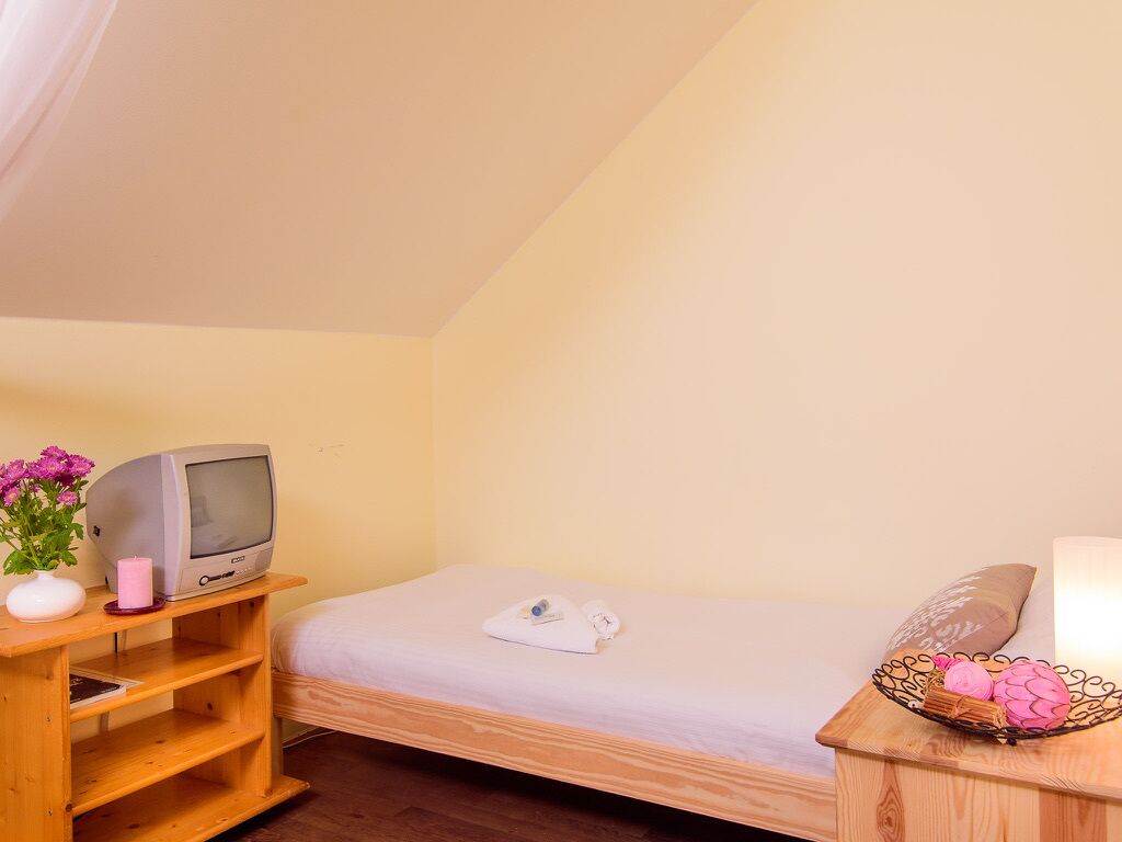 Ein kleines, gemütliches Zimmer mit einem Einzelbett, einem Nachttisch aus Holz mit Fernseher und einer rosa Kerze sowie einem Tisch mit einer Korb voller Snacks.