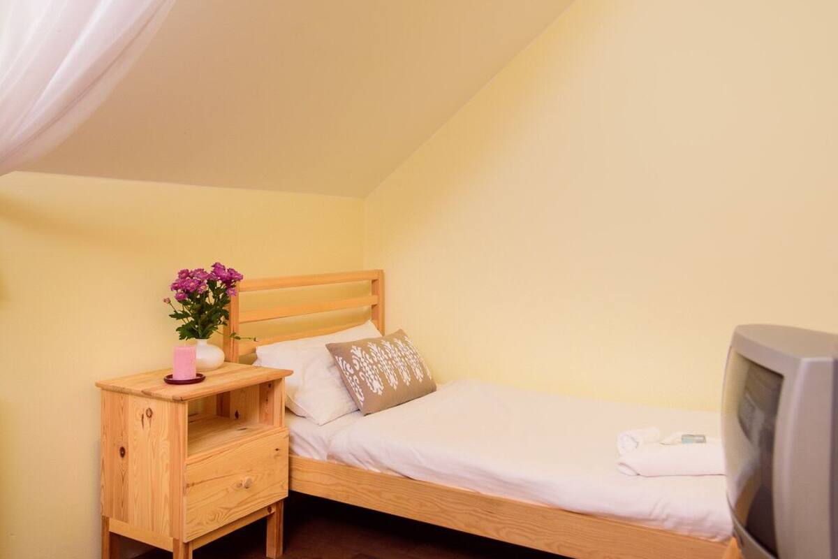Un lit simple avec une tête de lit en bois, une table de chevet avec une lampe et un téléviseur placé à droite.