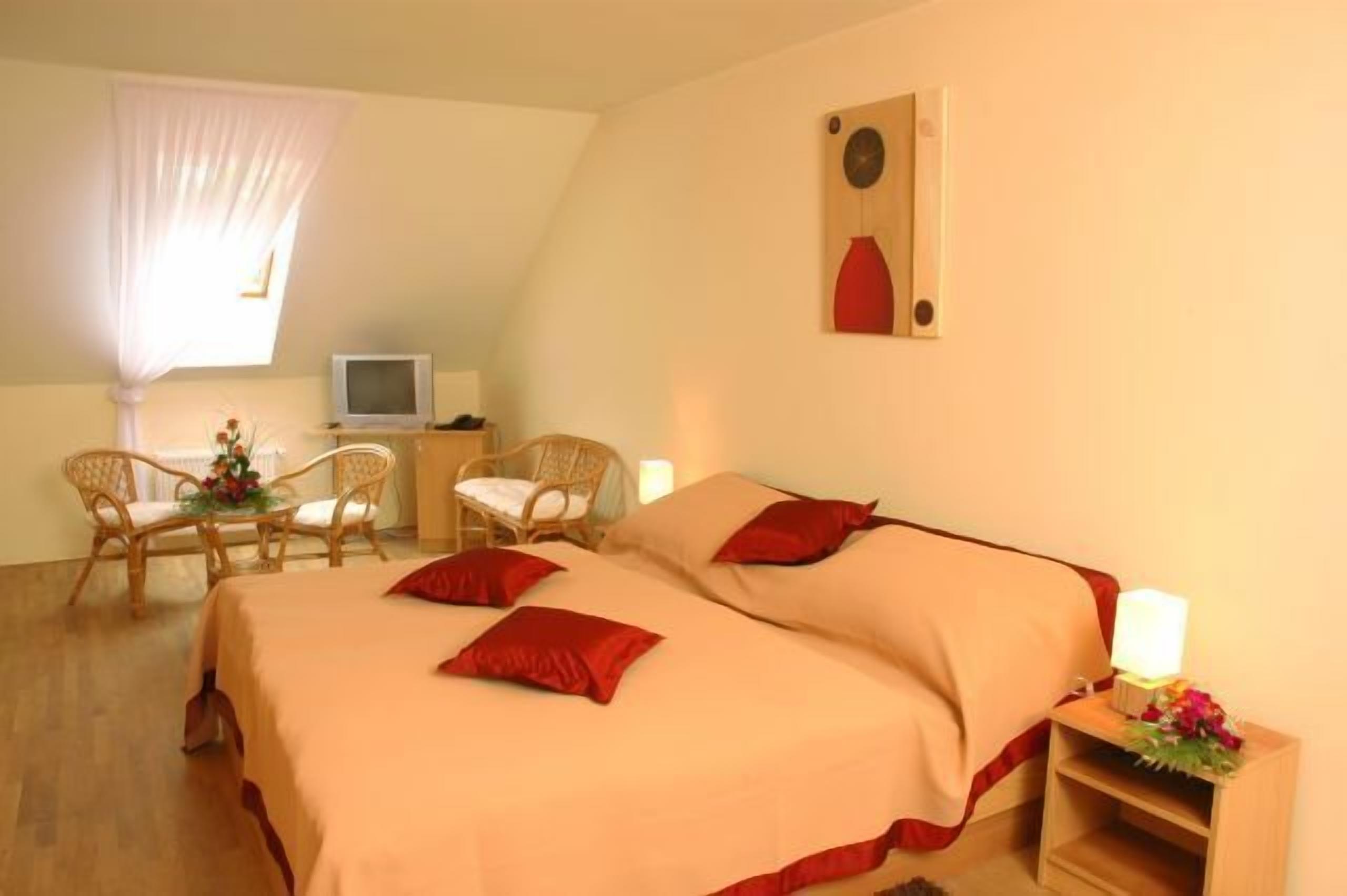 Een slaapkamer met een bed, twee stoelen, een klein tafeltje met bloemen, een televisie en een kunstwerk aan de muur.