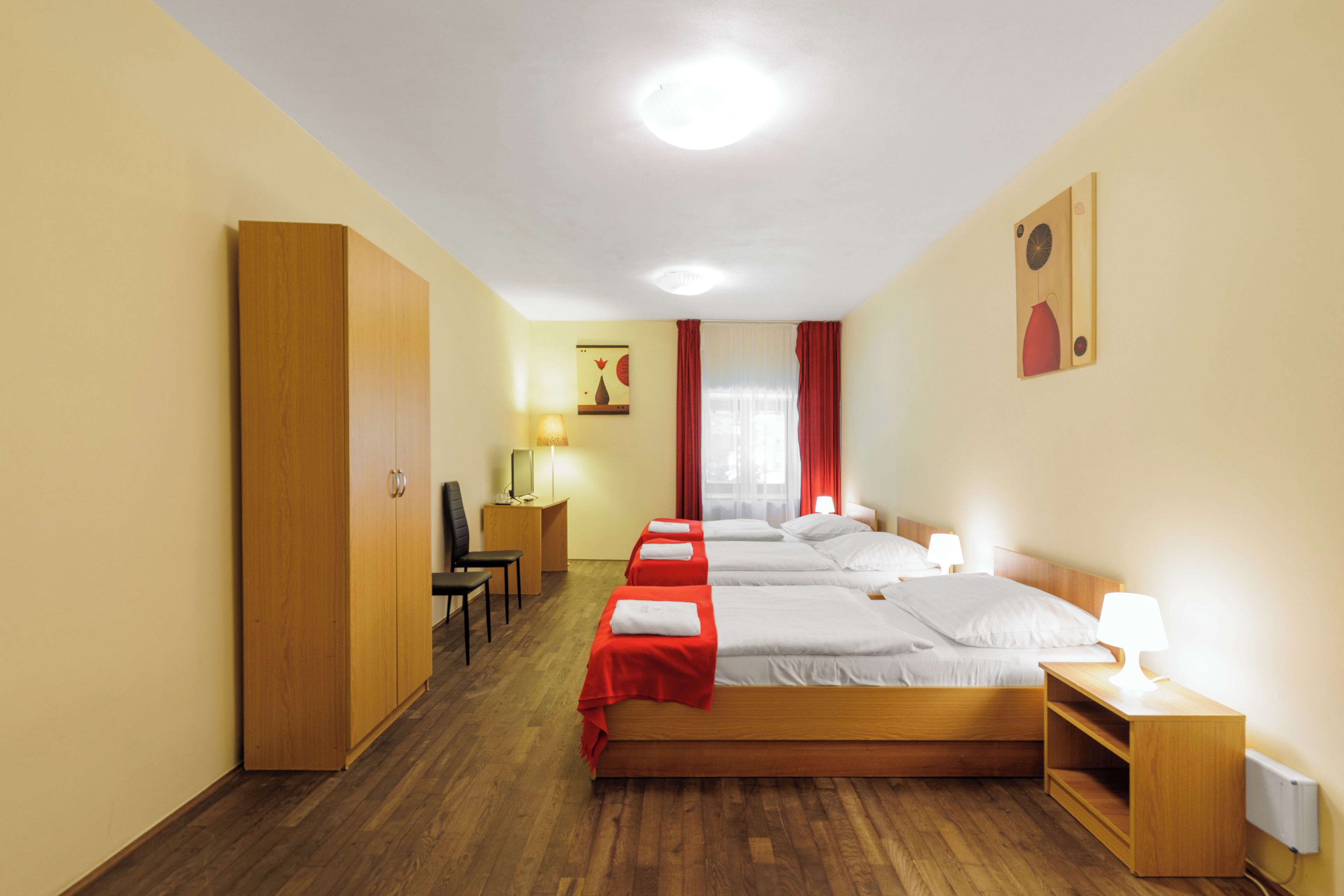 A hotel room with three beds, wooden wardrobe, desk, chair, and bedside lamps.