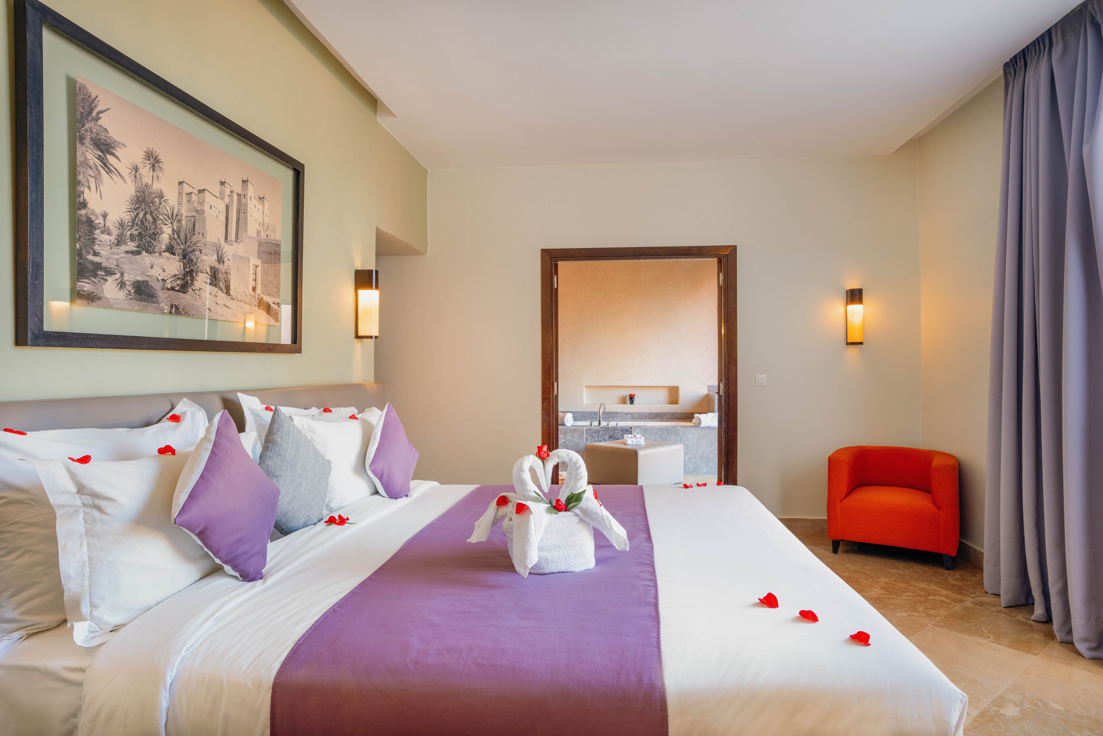 A hotel room with a large bed, a framed picture on the wall, a bathroom visible through an open door, and a red chair.