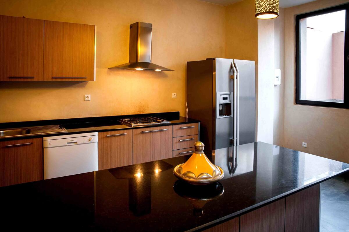 A modern kitchen with wooden cabinets, a stainless steel range hood, a refrigerator, and a black countertop.