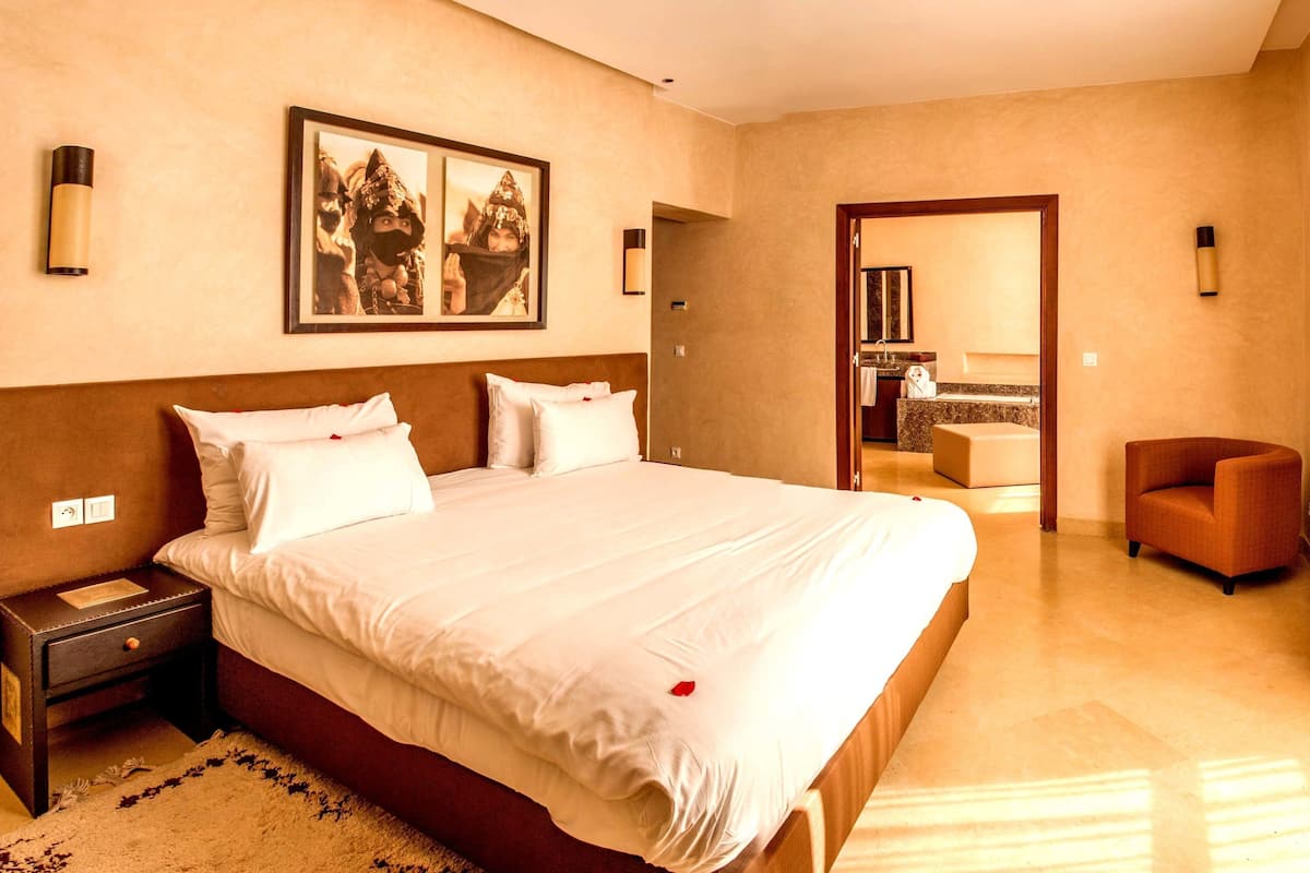 A hotel room with a large bed, a nightstand, a chair, a bathroom visible through an open door, and a framed picture on the wall.