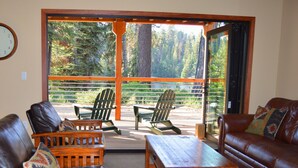 Una sala de estar acogedora con una mesa de centro de madera, dos sillones Adirondack y un sofá de cuero. Un ventanal ofrece vistas a un bosque y un lago.