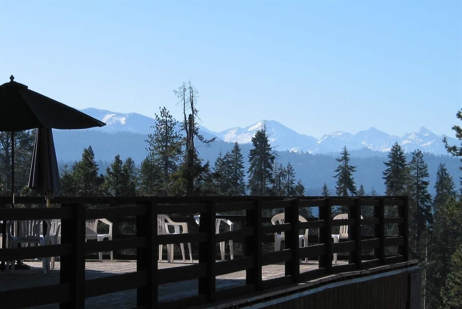 Eine Holzdielen-Terrasse mit Plastikstühlen, mit Blick auf eine gebirgige Landschaft mit schneebedeckten Gipfeln und dichtem Wald.