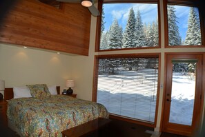 A bedroom with a large bed, a wooden ceiling, and a window with a view of snow-covered trees.