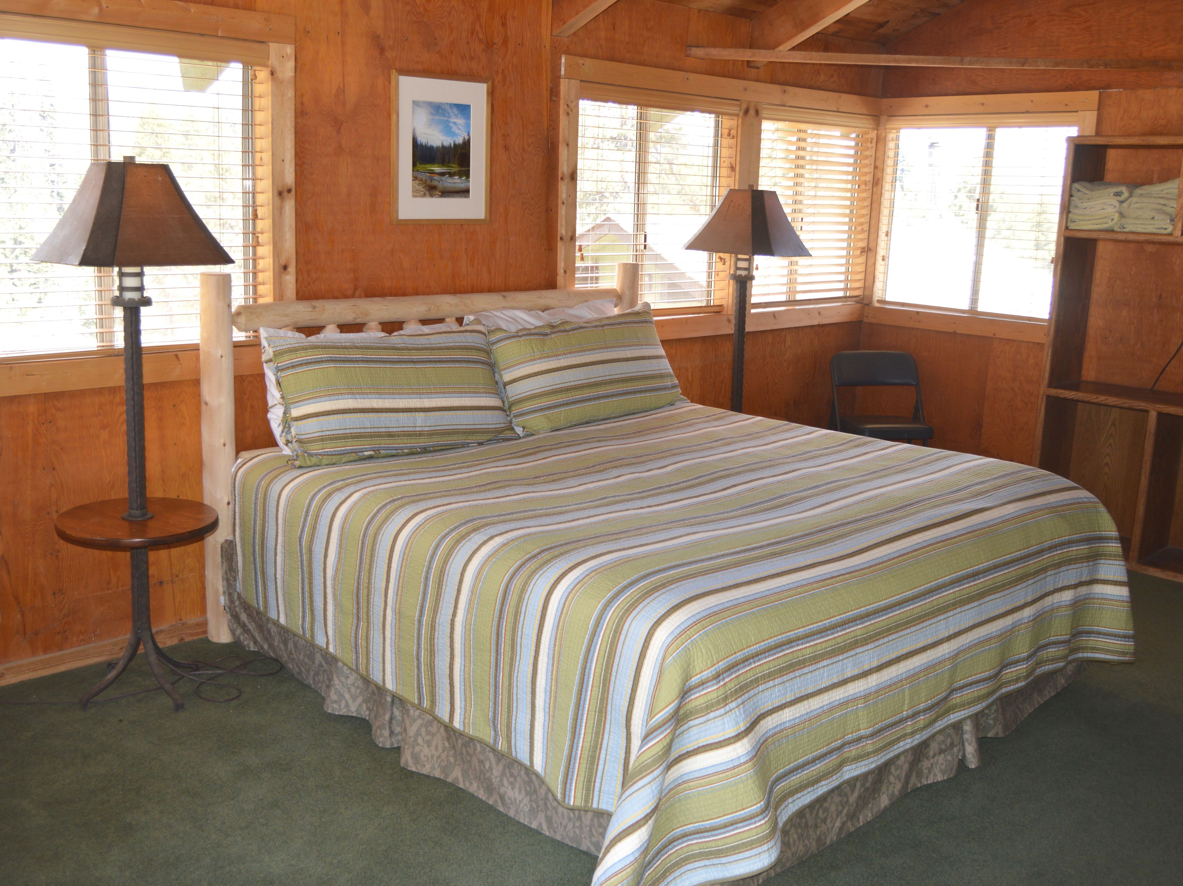 A wooden cabin bedroom with a bed, two floor lamps, a picture on the wall, and a window with blinds.