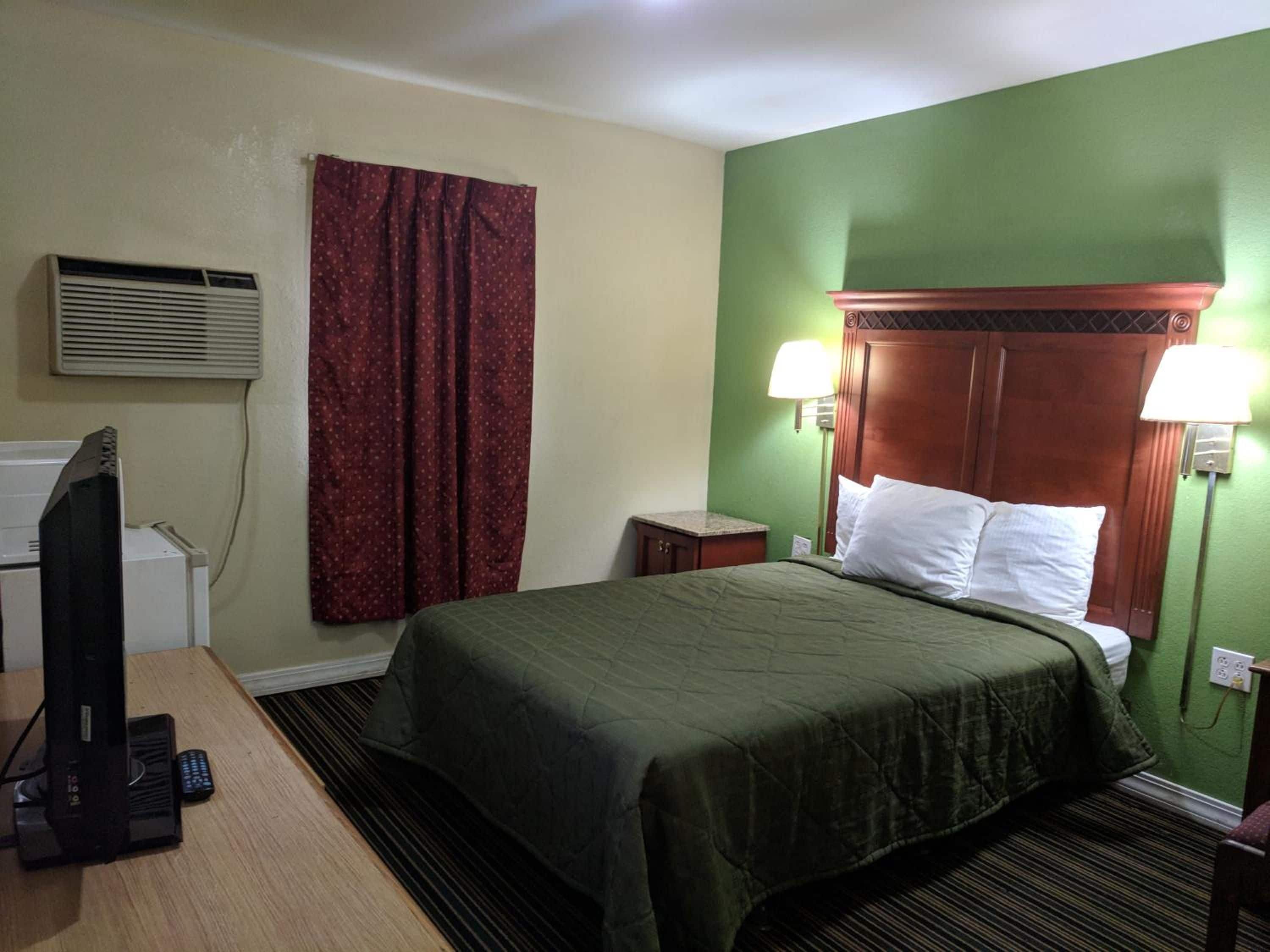 A hotel room with a bed, a desk with a television, an air conditioning unit, and a window with curtains.