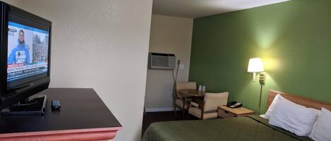 Une chambre d’hôtel comprenant un lit, un bureau avec une télévision, une table avec des chaises, une lampe et un appareil de climatisation.