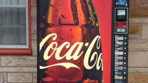 Een Coca-Cola automaat met een grote Coca-Cola afbeelding, een muntgleuf en een selectie drankjes waaronder Dasani, Mountain Dew en Pepsi.