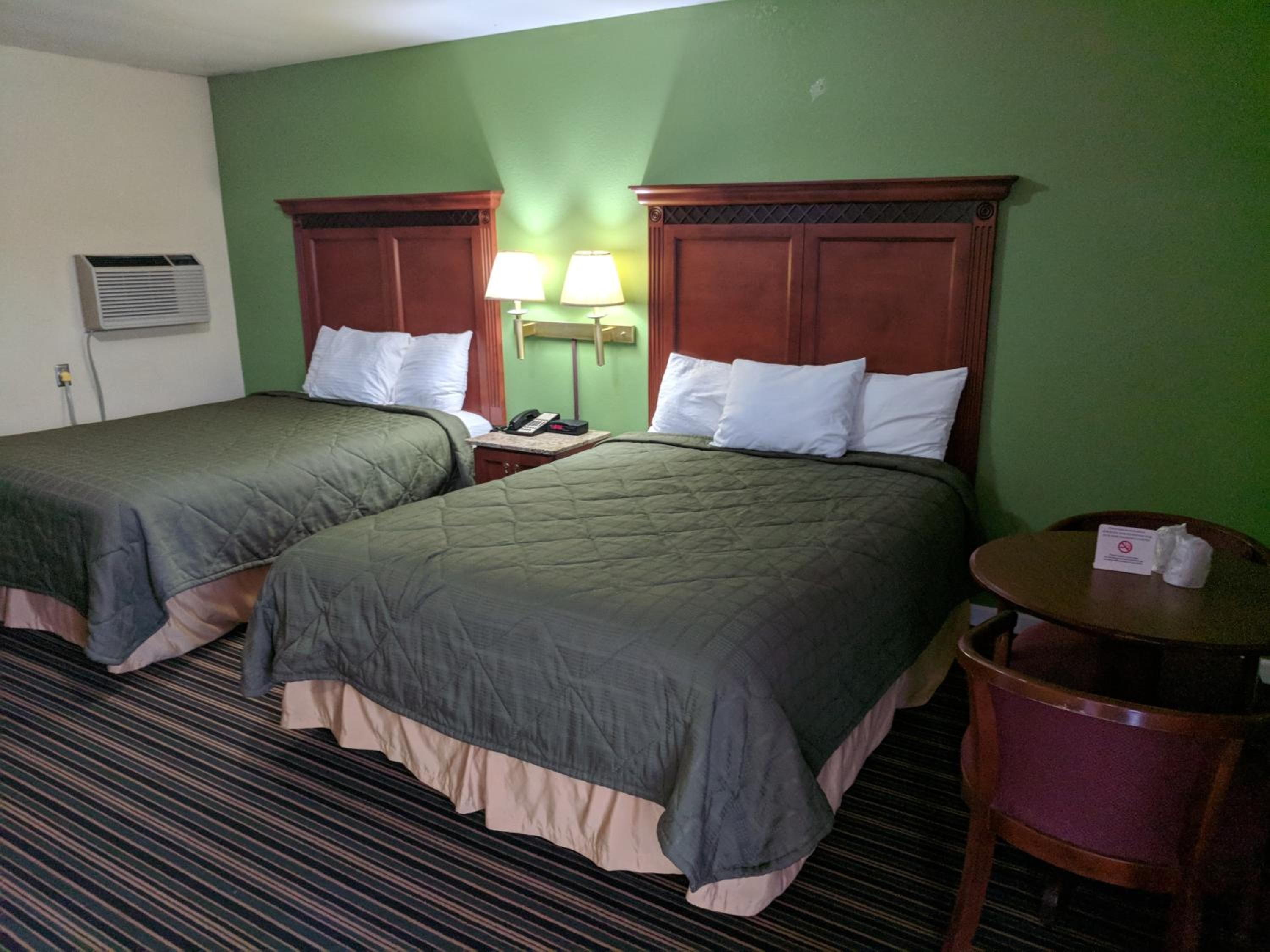 A hotel room with two beds, a bedside table with a lamp, a chair, and a small table with a tissue box.