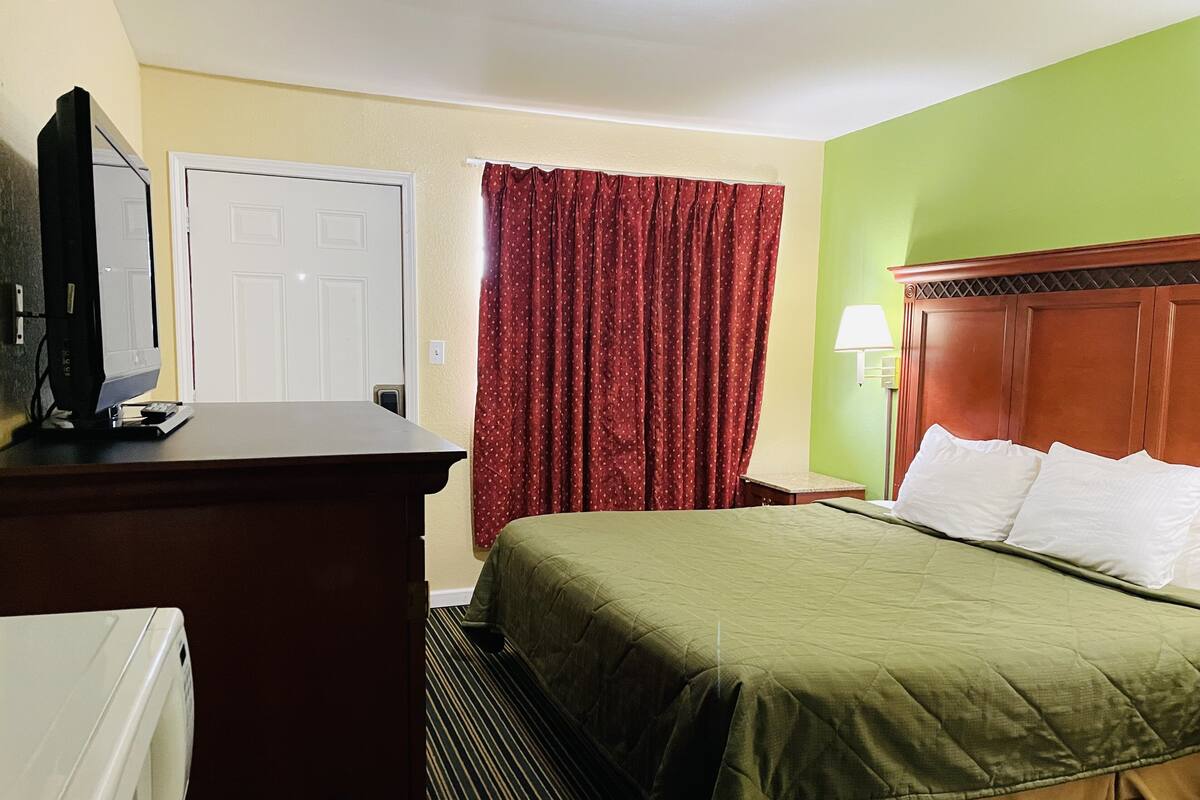 A hotel room with a bed, a desk with a television, a door, and a window with curtains.