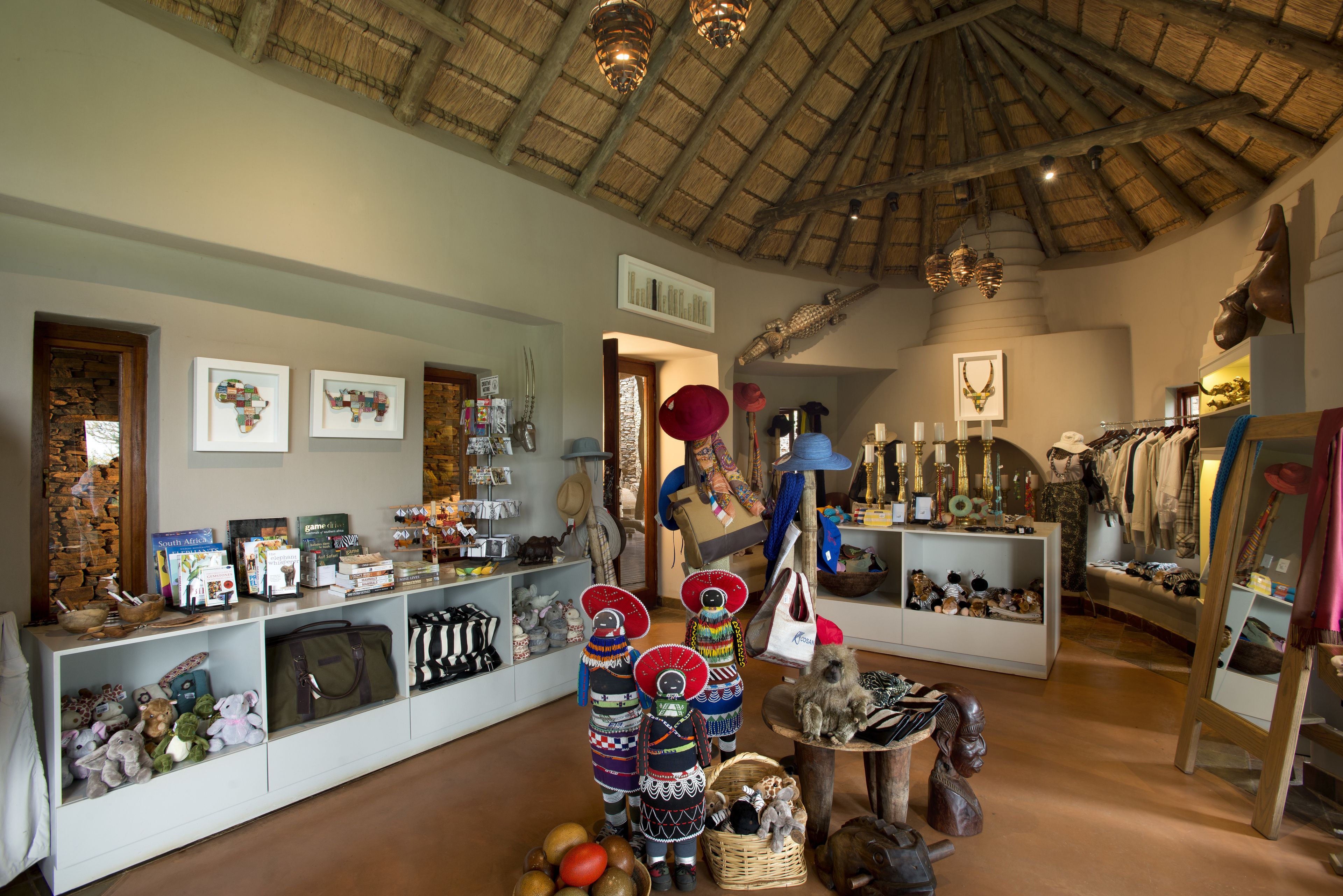 Interior de un hotel boutique con techo de paja, vigas de madera y una variedad de objetos expuestos, incluyendo textiles, bolsos y elementos decorativos.