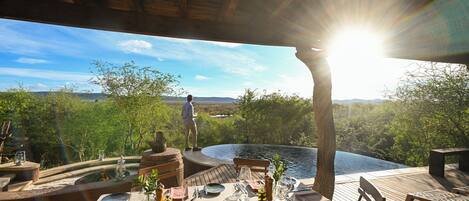 An outdoor dining area with a table set for two, overlooking a body of water and a mountainous landscape.