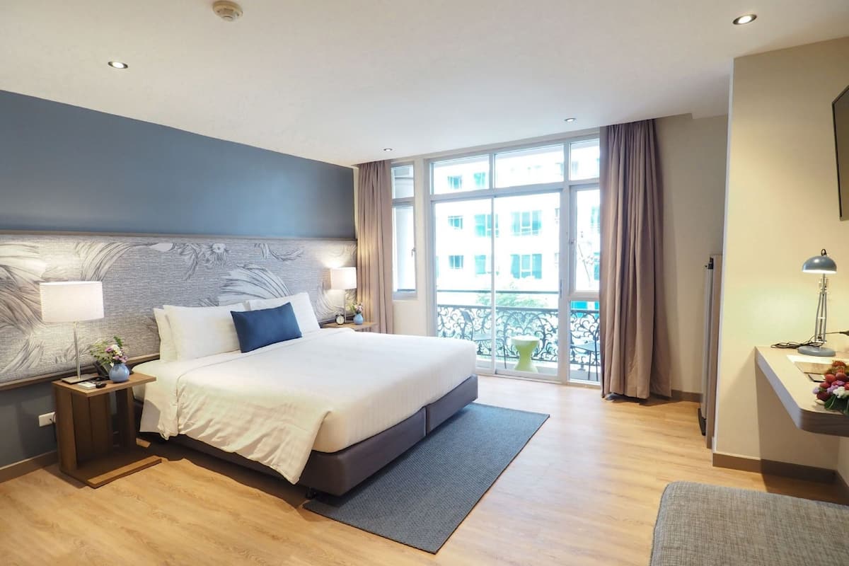 Une chambre d’hôtel avec un grand lit, une table de chevet avec une lampe, un bureau avec une lampe et une vue sur les bâtiments par la fenêtre.