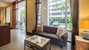 Un moderno vestíbulo de hotel con un mostrador de recepción, un sofá con cojines, una mesa de centro con revistas y grandes ventanas que ofrecen vistas a la calle y edificios cercanos.
