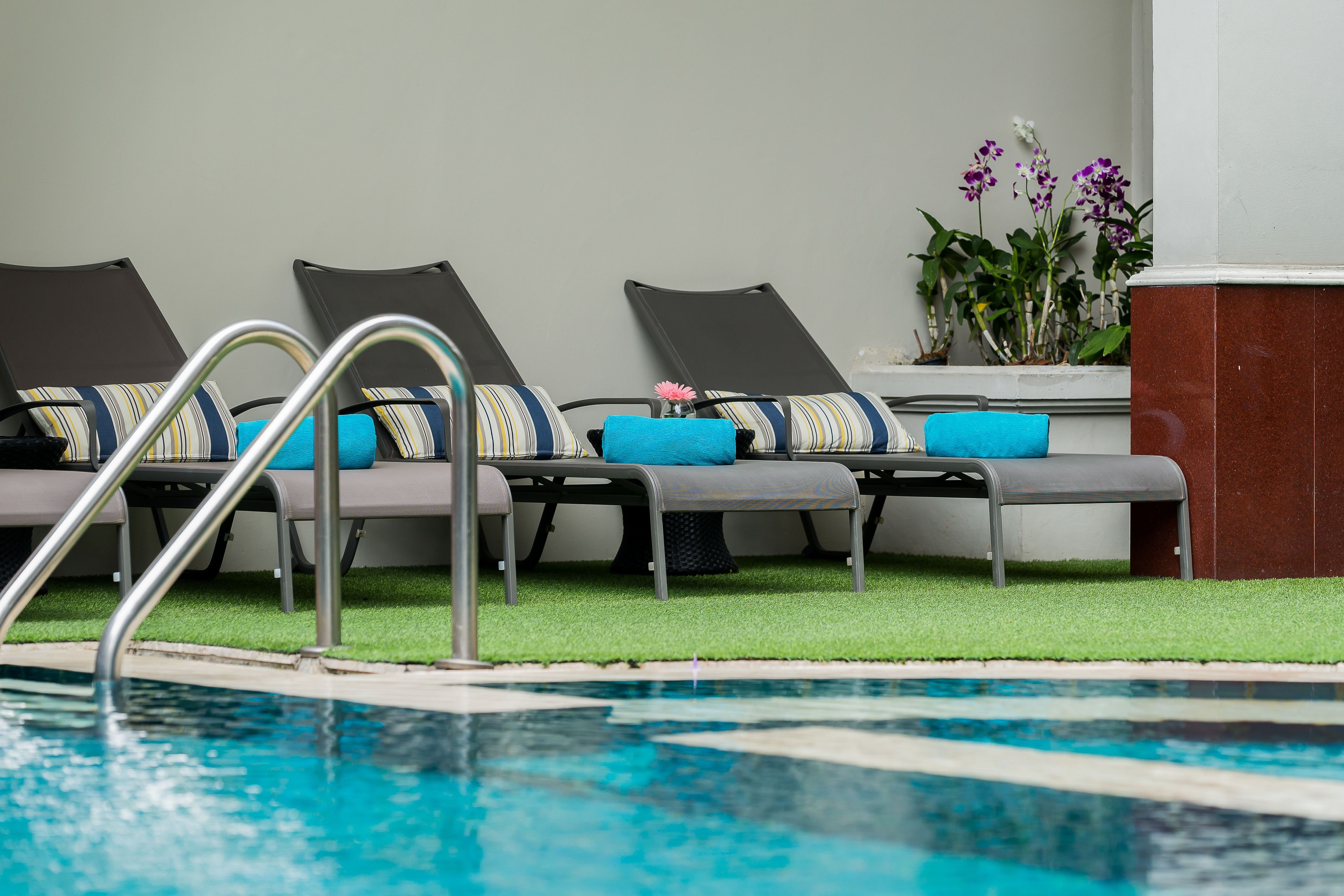 Un espace aménagé au bord de la piscine, avec des chaises longues, un escalier métallique pour accéder à la piscine et du gazon synthétique.