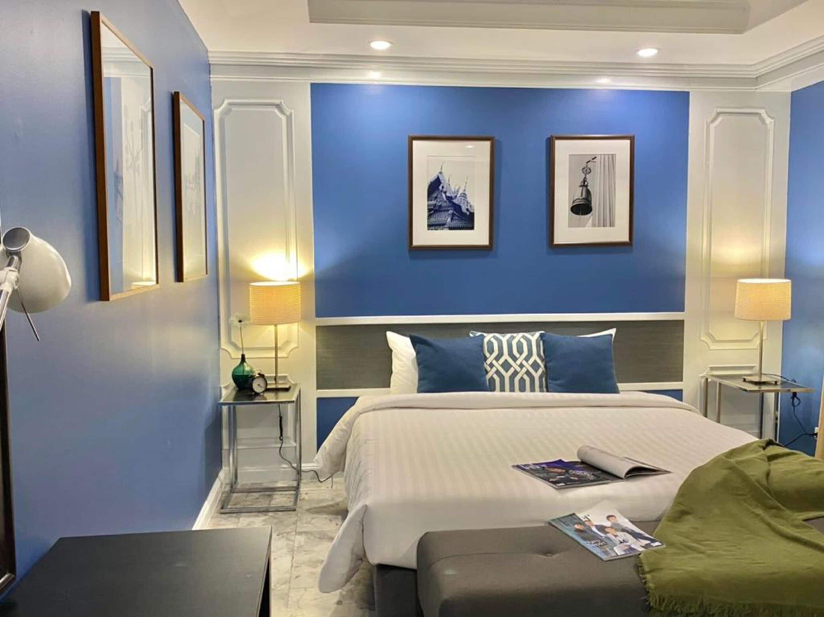 A modern hotel room with a large bed, two bedside tables with lamps, a green armchair, and framed artwork on the walls.